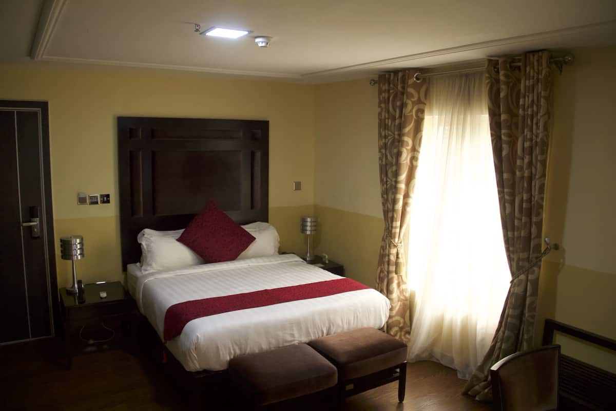 A hotel room with a large bed, a bedside table with a lamp, a chair, and a window with curtains.