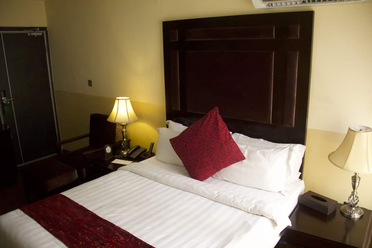 A hotel room with a bed, a nightstand, a lamp, a clock, and a door.