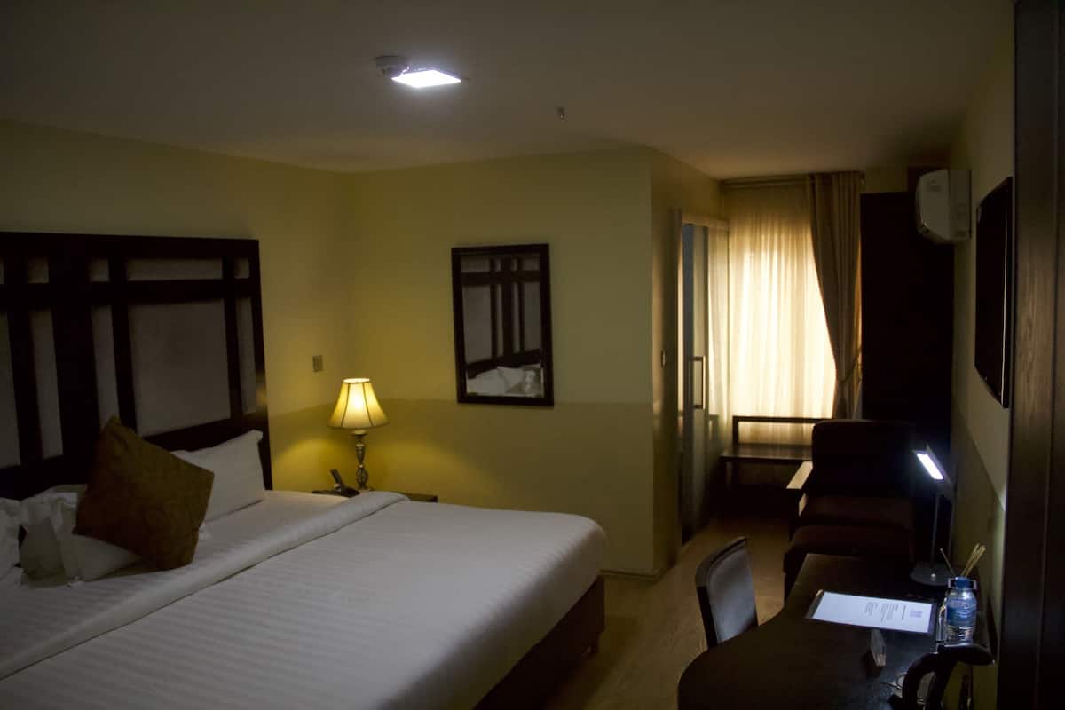 A hotel room with a bed, a desk with a chair, a lamp, and a window with curtains.