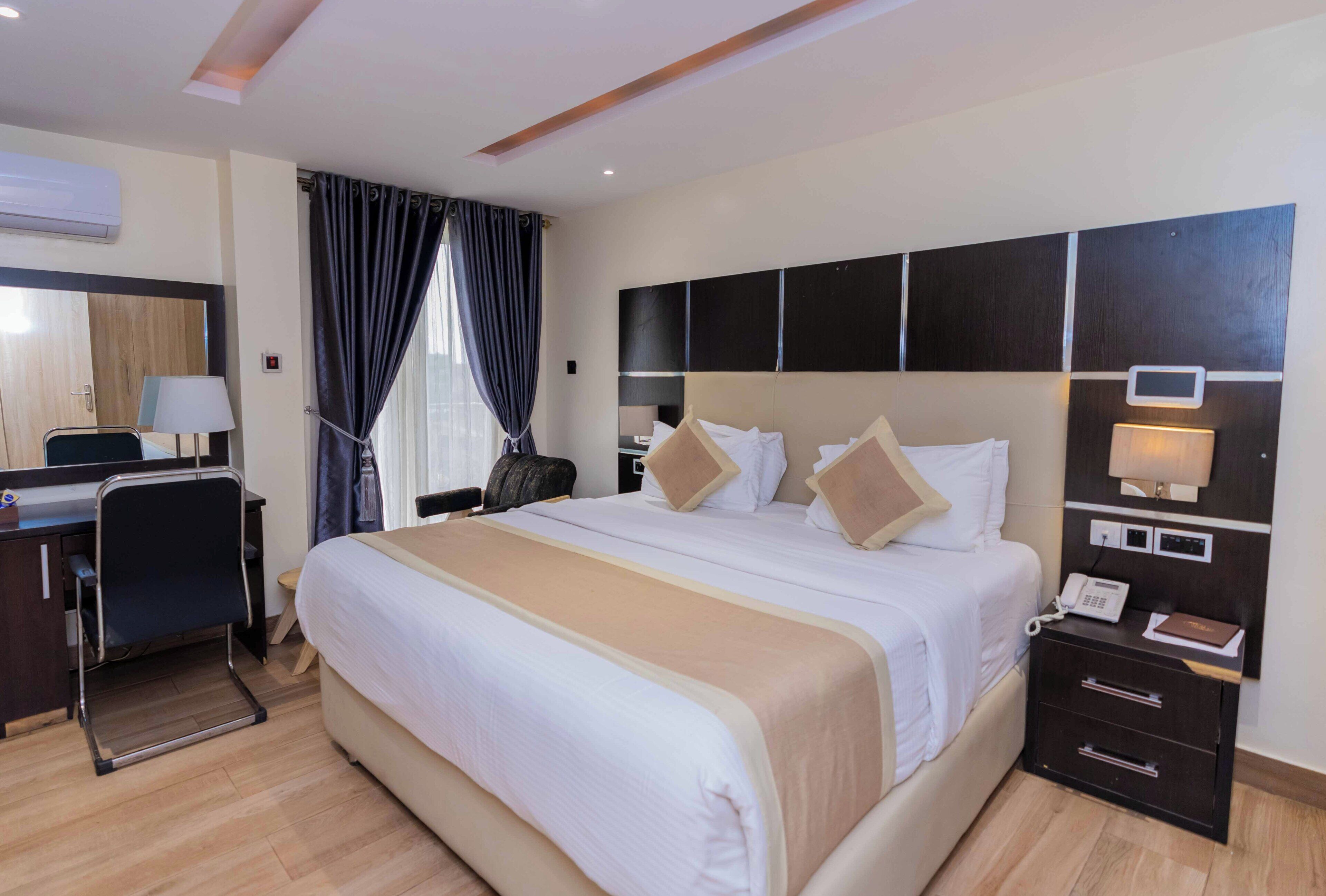 A hotel room with a large bed, a desk with a chair, a bedside table with a lamp, and a wall-mounted TV.