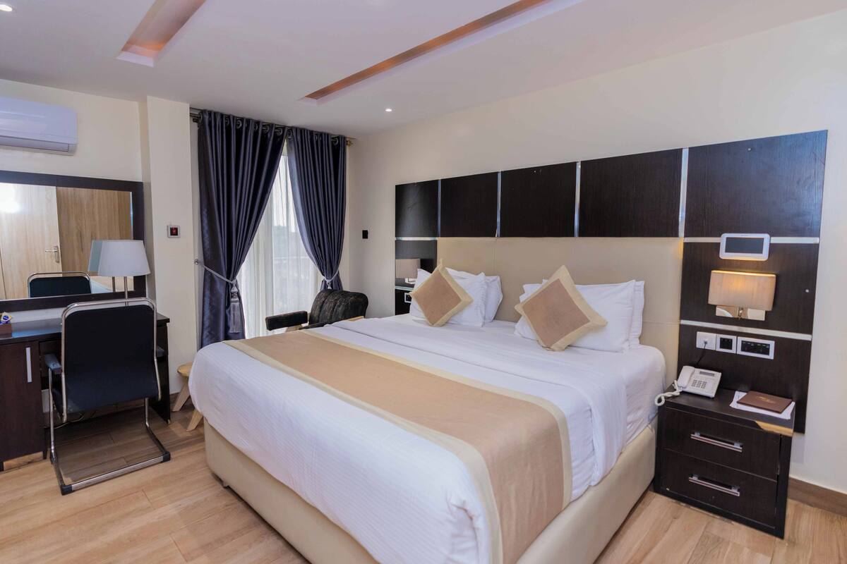 Quarto de hotel com uma cama grande, uma escrivaninha com cadeira, mesa de cabeceira com abajur e uma televisão na parede.