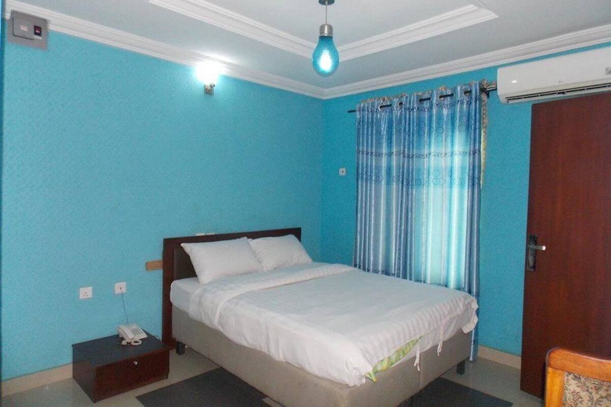 Quarto com cama, mesa de cabeceira com telefone, parede azul, janela com cortinas e ar condicionado.