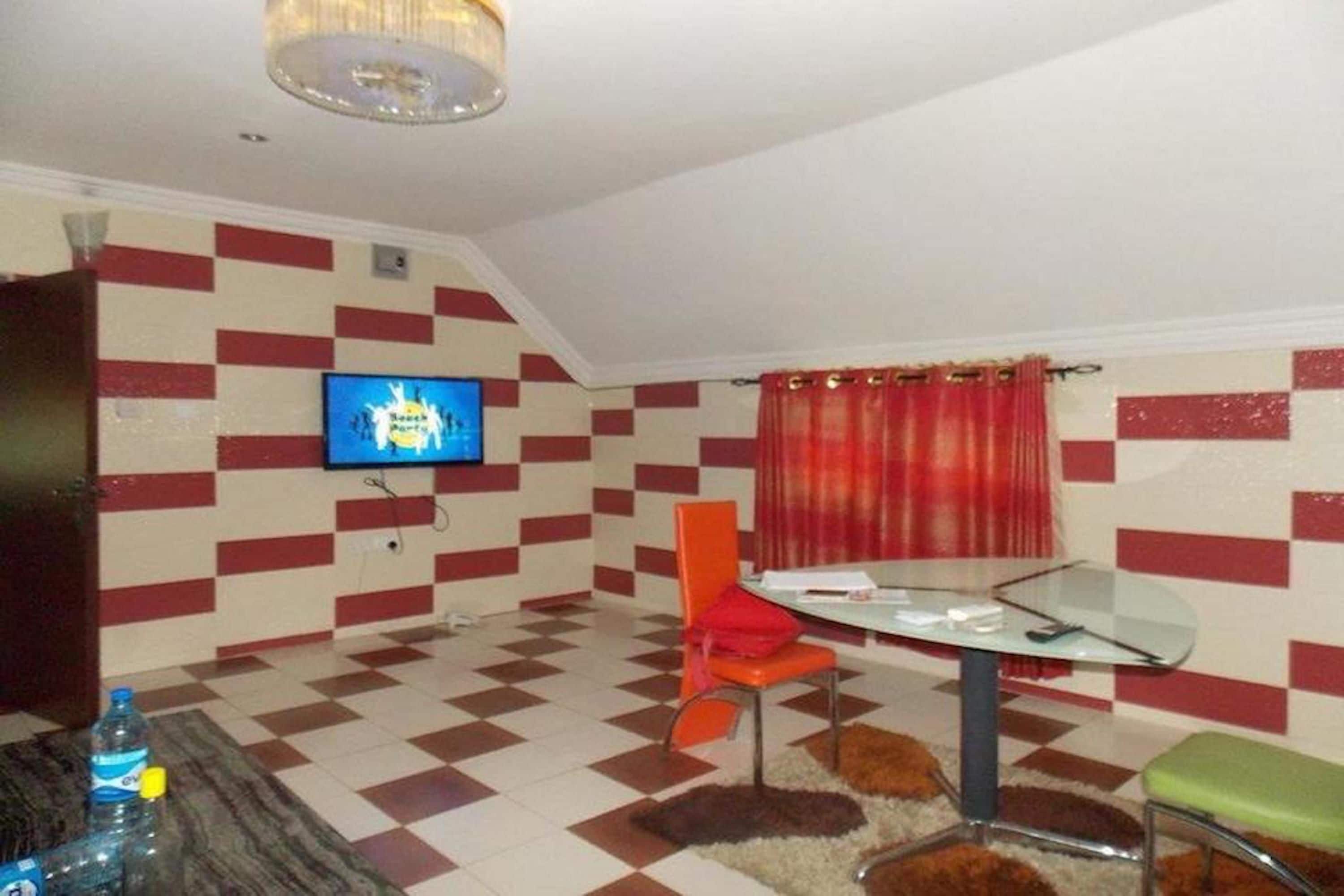Een ruimtelijk ingerichte leefruimte met een zwart-wit betegelde vloer, een aan de muur bevestigde televisie, een eettafel met stoelen en een klein vloerkleed.