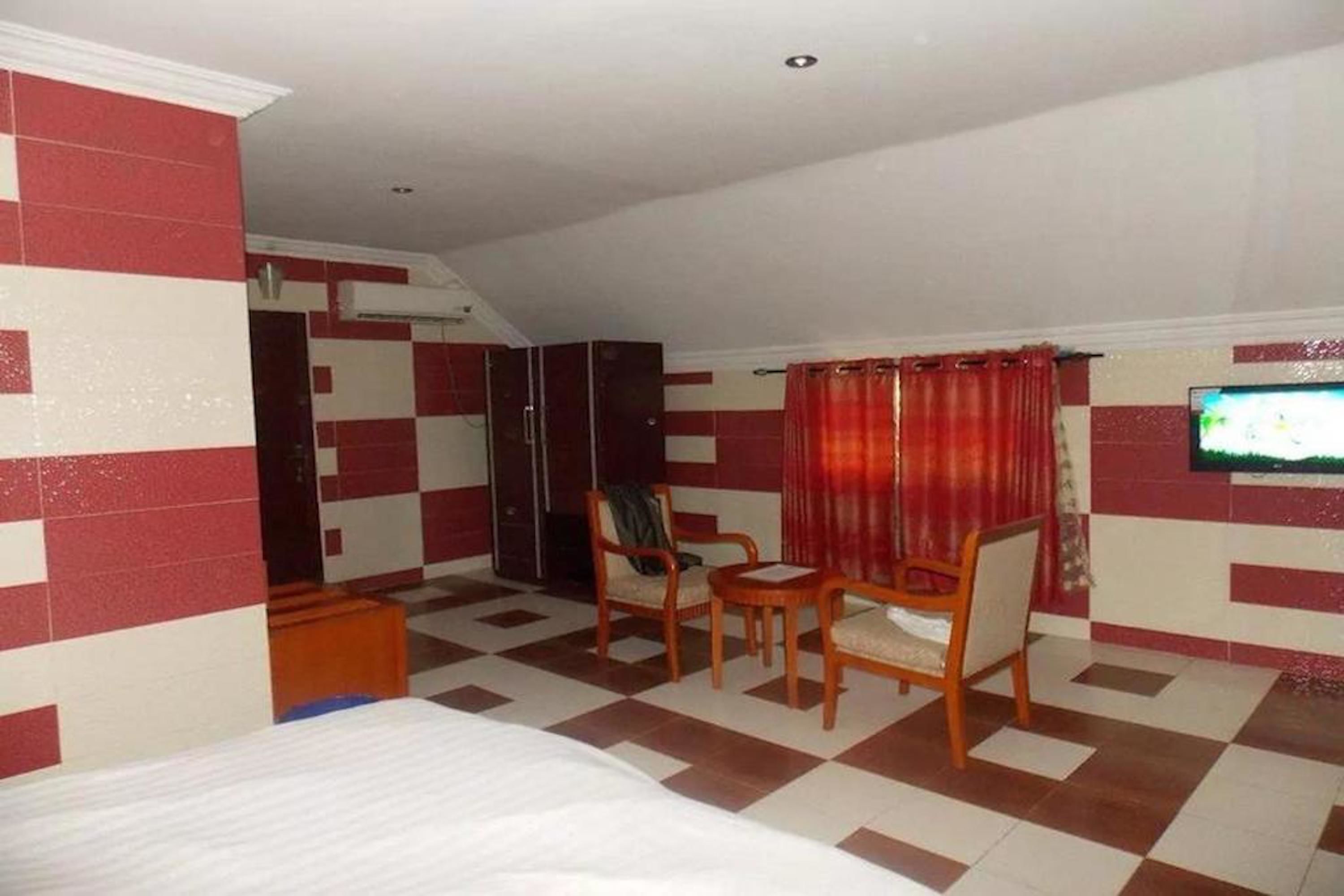 Quarto de hotel com piso quadriculado, uma cama, uma televisão e um espaço de estar com cadeiras.