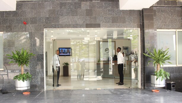 A reception area with a glass door, potted plants, a television screen, and a push button sign. - Pelican Hotel Lekki (Lekki)