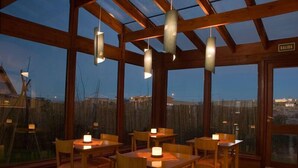 A restaurant with wooden tables and chairs, hanging pendant lights, and large windows offering a view of the outside. - Hosteria Roble Sur (El Calafate)