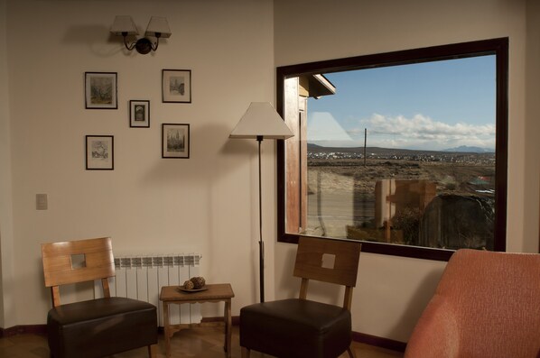 Una habitación con dos sillas de madera, una mesita con adornos, una lámpara de pie y un amplio ventanal con vistas a un paisaje lejano.
