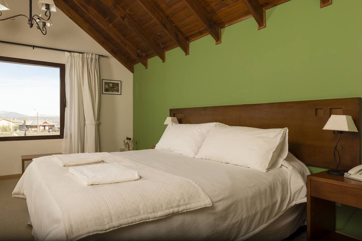 Una cama bien hecha con sábanas blancas, cabecera de madera y mesita de noche con lámpara y teléfono.