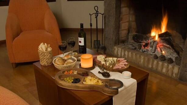 A cozy room with a fireplace, a wooden table set with food and drinks, and an orange armchair. - Hosteria Roble Sur (El Calafate)