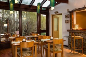 A restaurant with wooden tables and chairs, a stone bar, and a view of trees through large windows. - Hosteria Roble Sur (El Calafate)