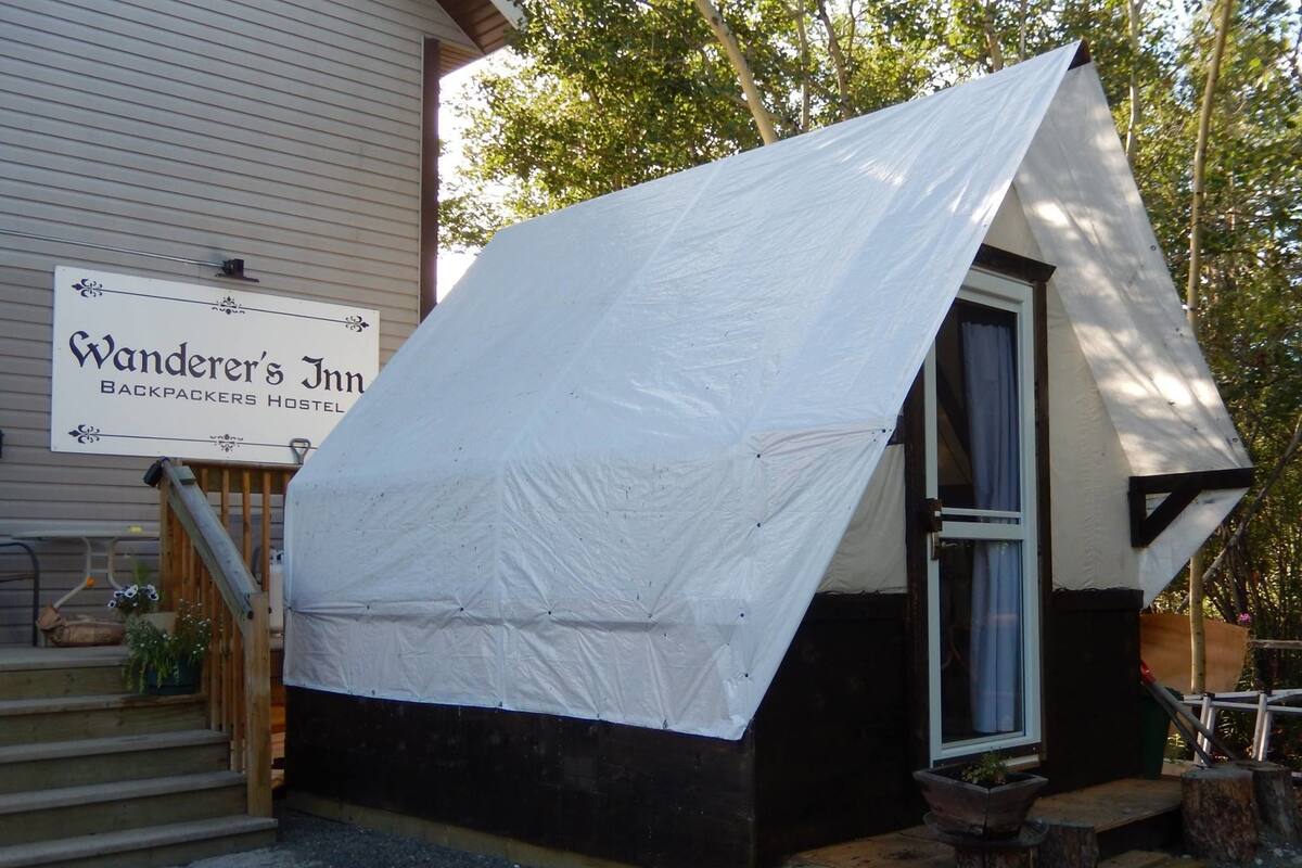A small, single-story building with a white tent-like structure on the roof, a wooden door, and a sign indicating it is a backpackers hostel.