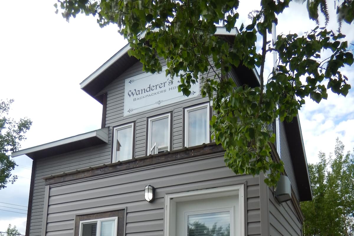 A small, single-story building with a sign reading "Vander's Backpackers House." It features a front door, a small window, and a wooden bucket near the entrance.