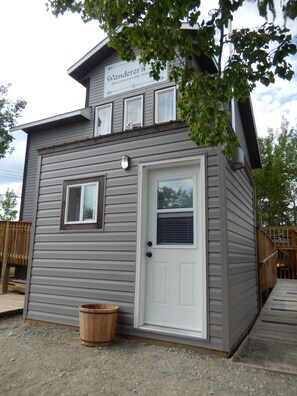 A small, single-story building with a sign reading "Vander's Backpackers House." It features a front door, a small window, and a wooden bucket near the entrance. - Wanderer's Inn Backpackers Hostel (Haines Junction)