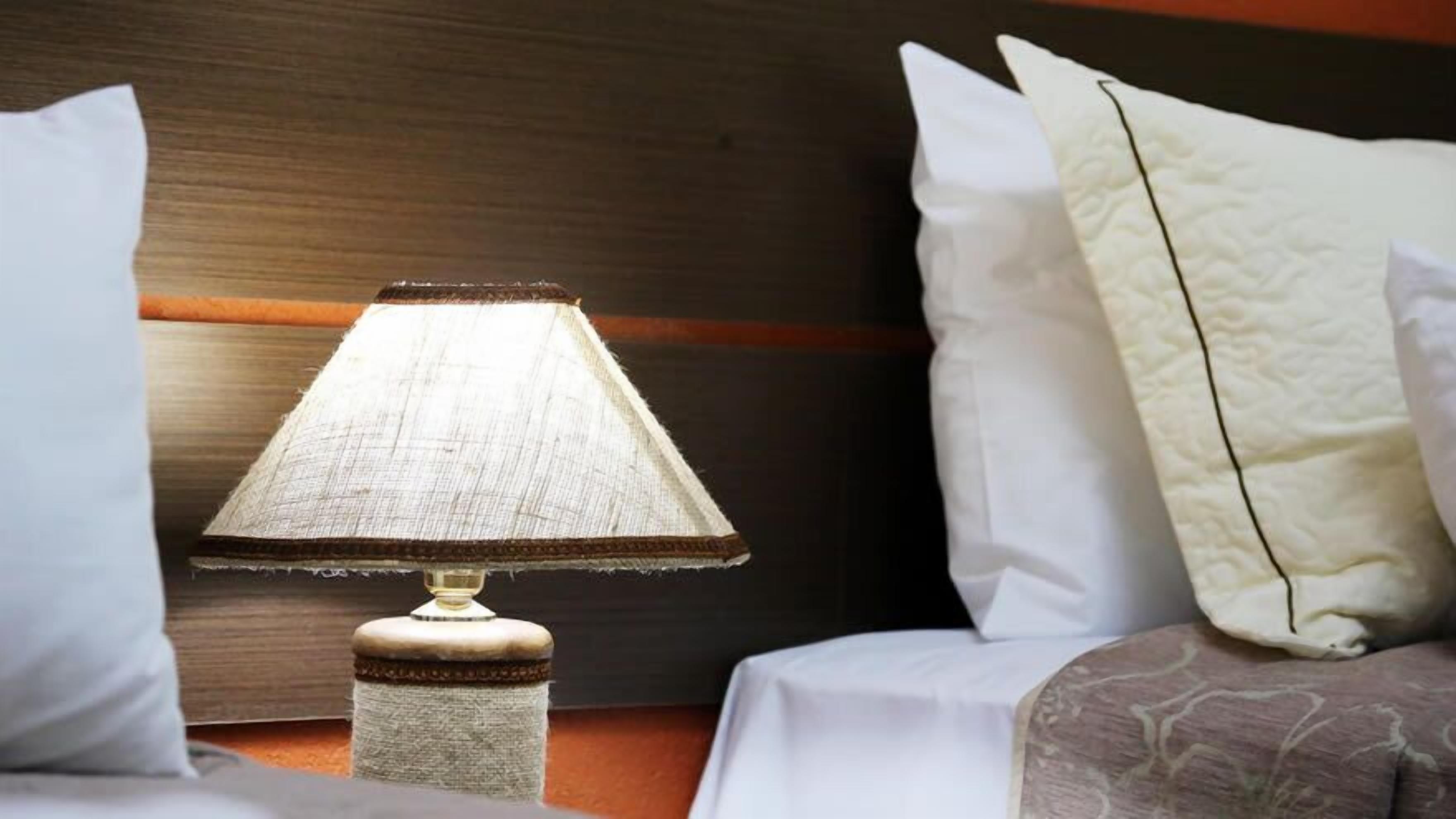 A bed with white bedding, a textured lamp on a wooden nightstand, and a dark wooden headboard.