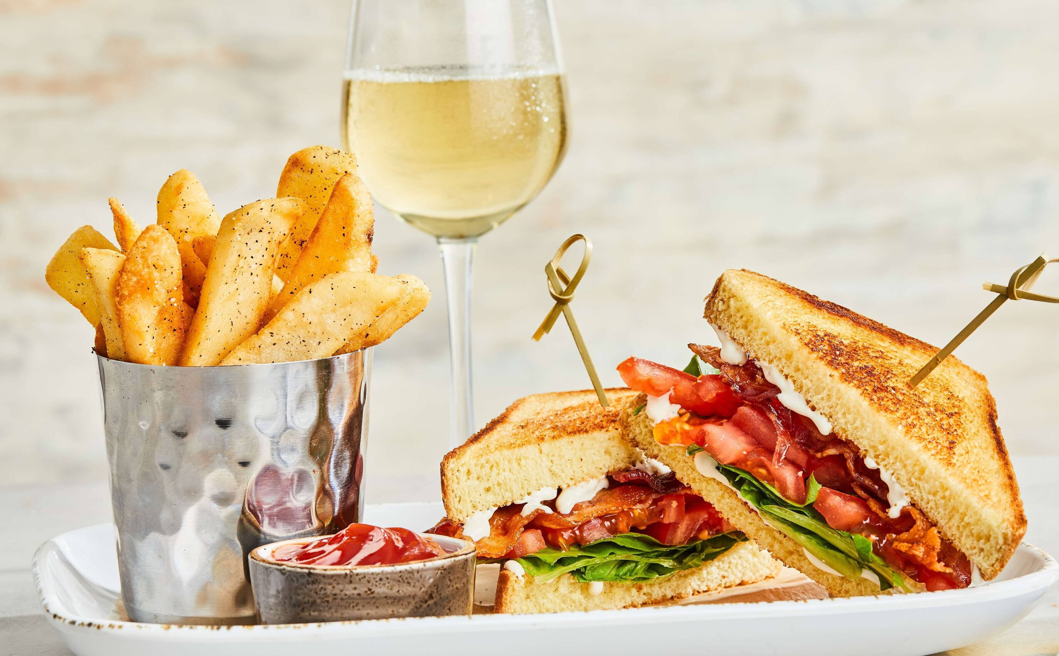 Un sandwich accompagné d’une portion de frites et d’un verre de vin blanc.
