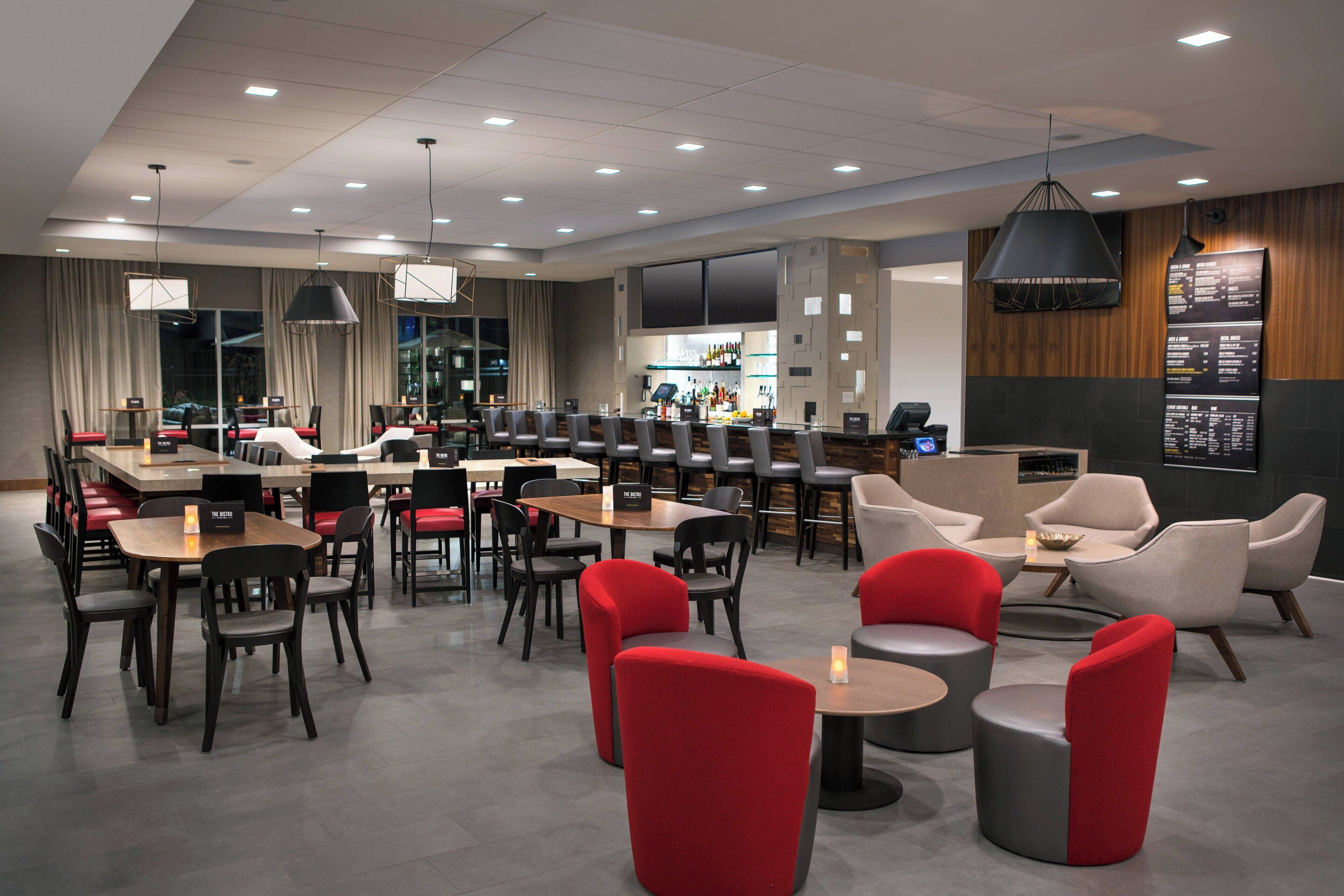 A modern hotel lobby with multiple seating areas, including tables and chairs, and a bar area with a menu display.