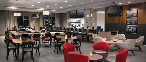 A modern hotel lobby with multiple seating areas, including tables and chairs, and a bar area with a menu display.
