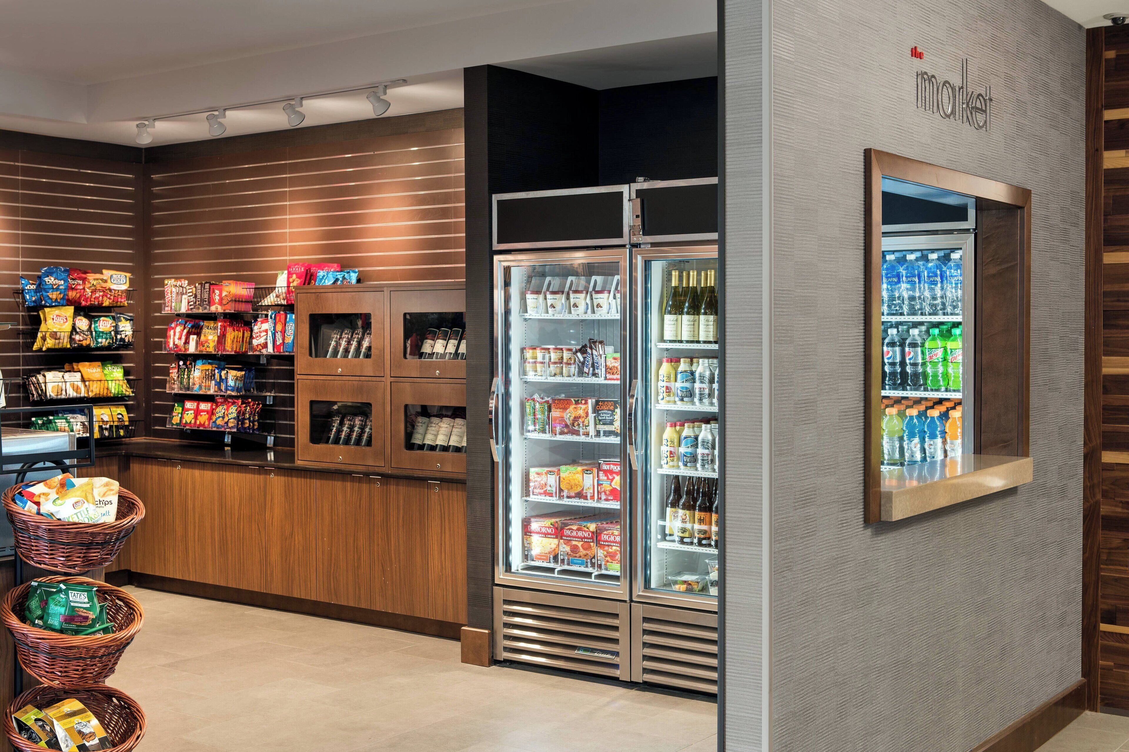 Interior de una tienda de comestibles con estantes llenos de snacks, una sección refrigerada con bebidas y un mostrador de madera con cestas.