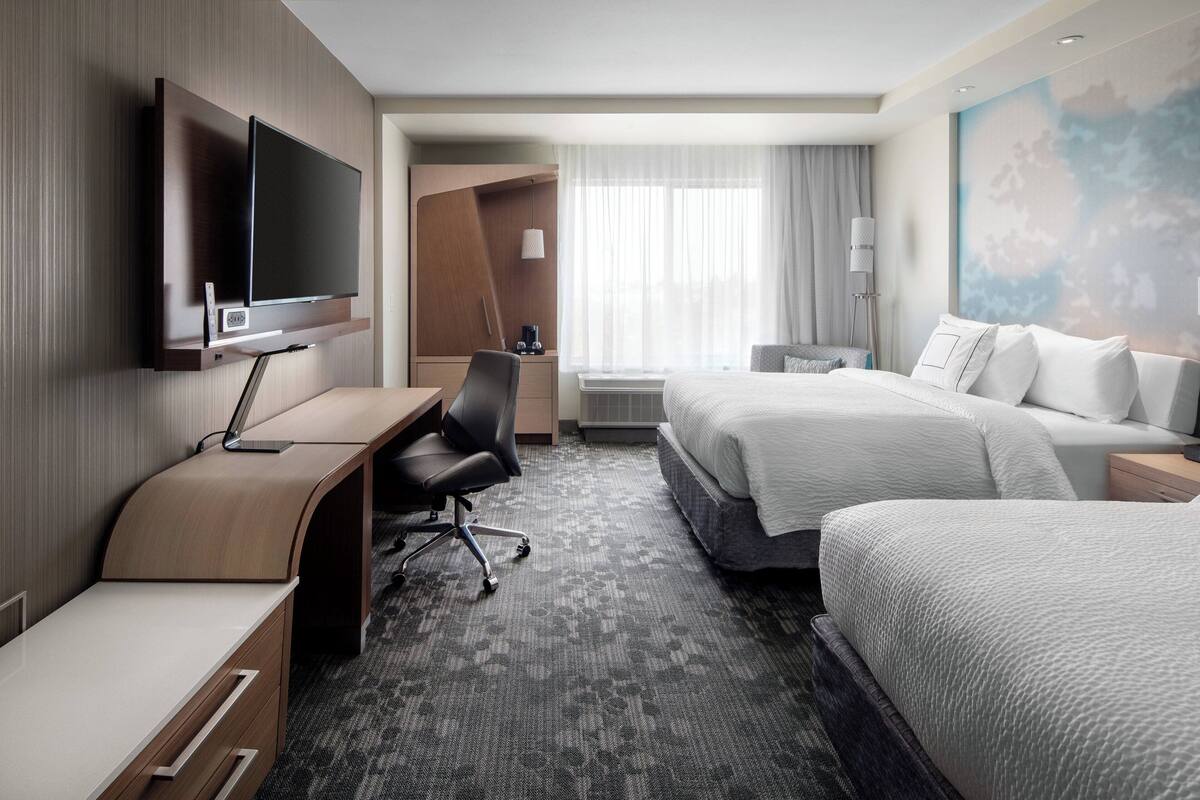 A hotel room with a flat-screen TV mounted on the wall, a desk with a chair, a bed with white bedding, and a large window with sheer curtains.