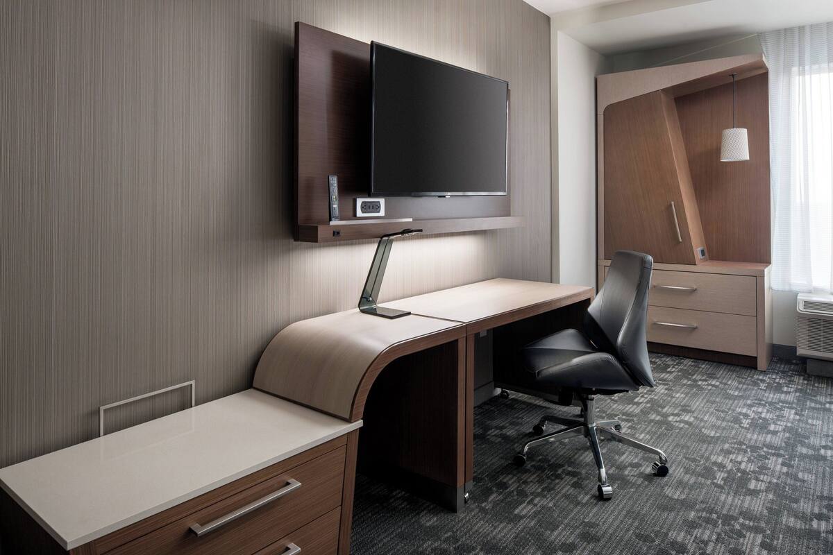 A hotel room with a flat-screen TV mounted on the wall, a desk with a chair, and a built-in wardrobe.
