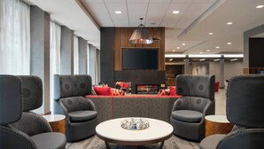 A modern lounge area with a round table, grey upholstered chairs, a flat-screen TV mounted on the wall, and a fireplace with red cushions.