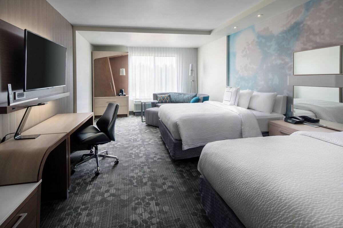 A hotel room with a flat-screen TV, a desk, two beds, a wardrobe, and a large window with curtains.