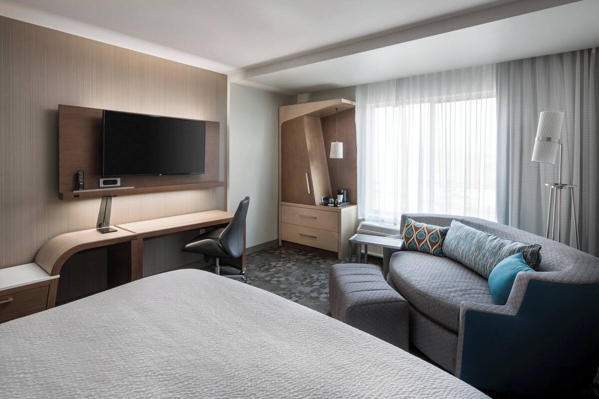 A hotel room with a bed, a desk with a flat-screen TV, a sofa, a desk lamp, and a window with curtains.
