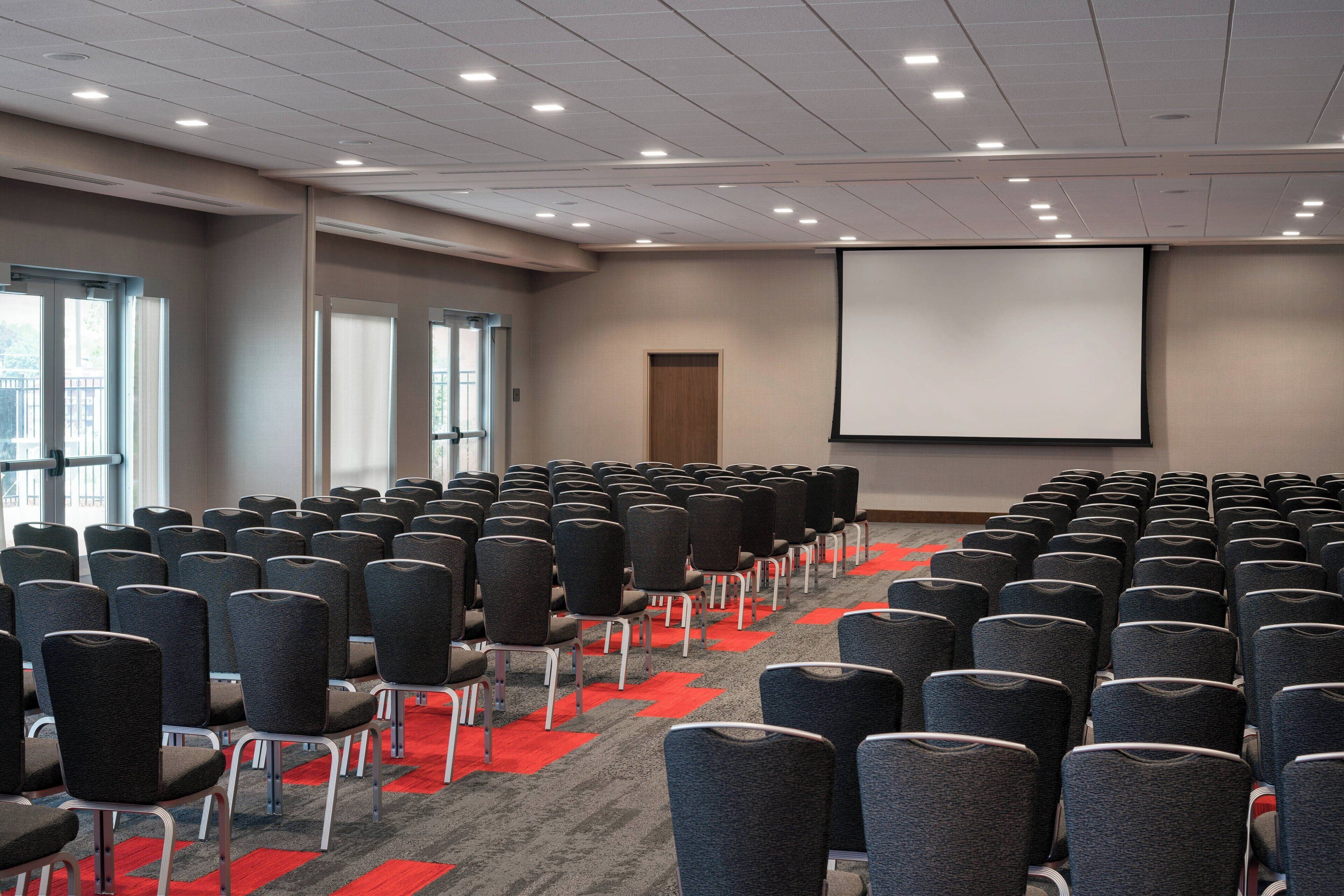 A conference room with rows of chairs, a large screen, and windows with blinds.