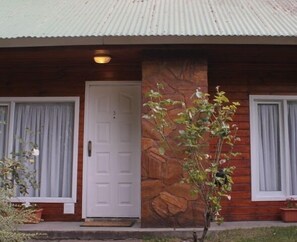 A wooden house with a stone pillar, a white door, and two windows with curtains. - Los Charitos (El Calafate)