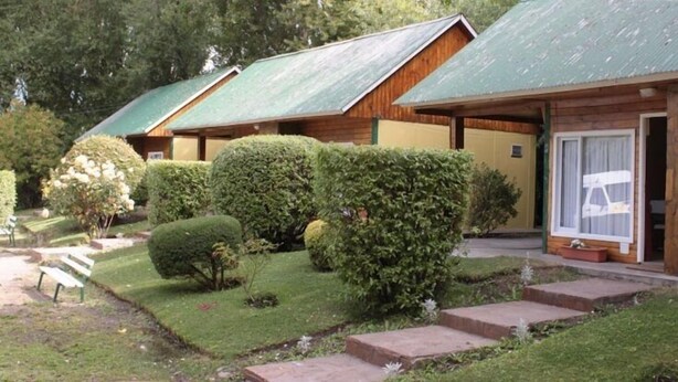 A row of wooden cabins with green roofs, surrounded by well-maintained gardens and a paved pathway. - Los Charitos (El Calafate)