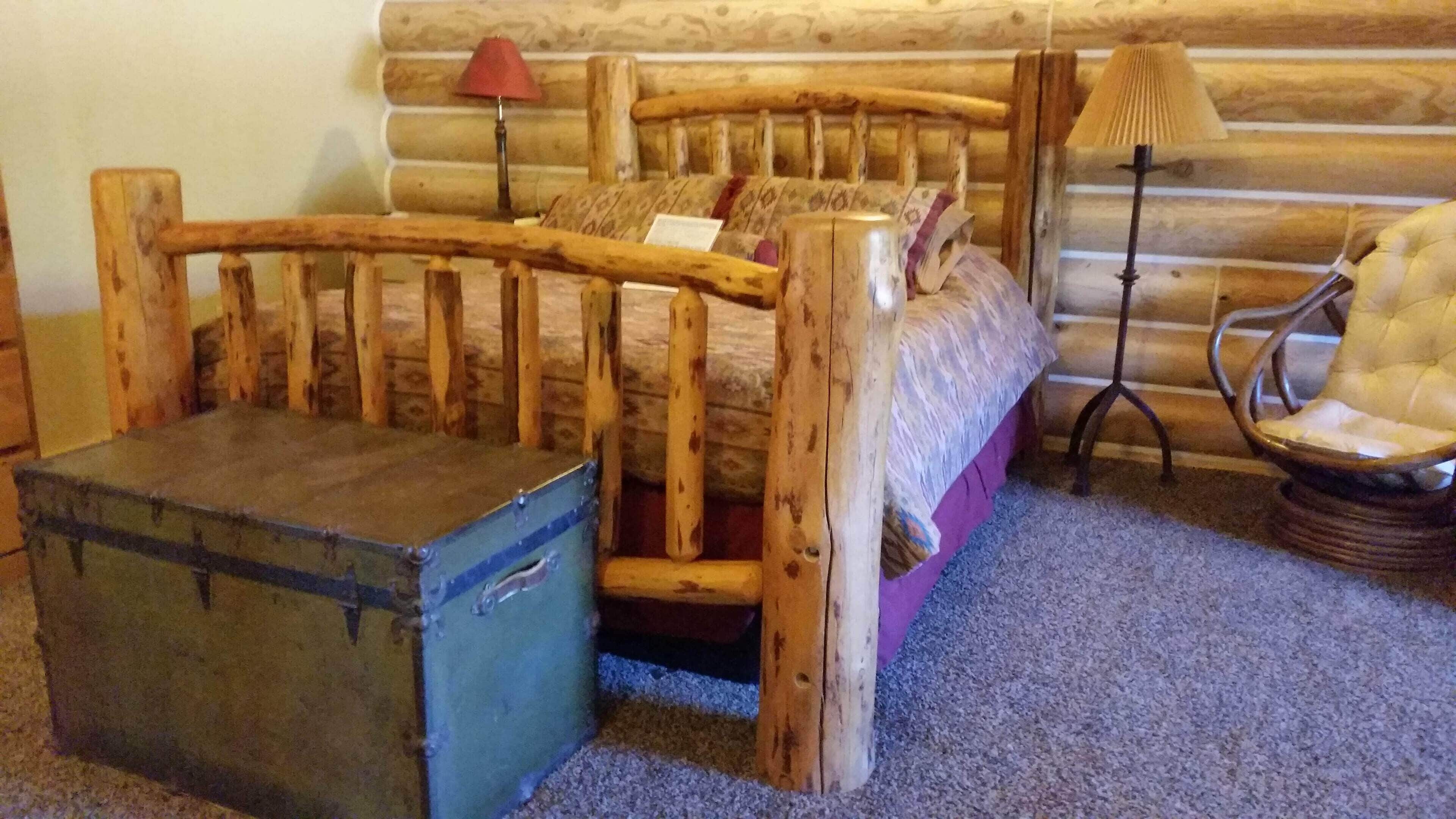 A rustic wooden bed with a headboard, a small chest of drawers, a chair, and a lamp.