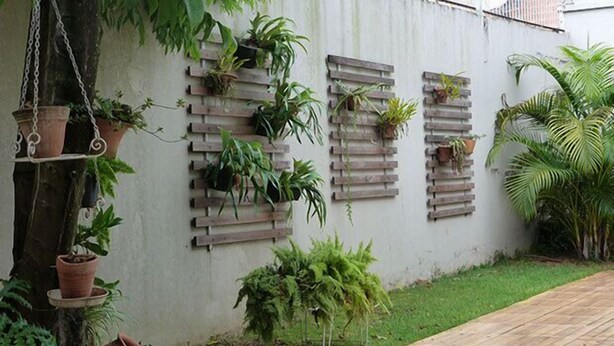 Un jardin avec des plantes en pot disposées le long d’un mur, une terrasse en bois et une zone herbeuse.