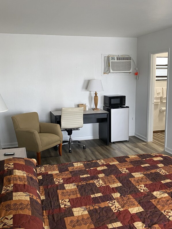 A hotel room with a bed, a desk, a chair, a lamp, a microwave, and an air conditioning unit.