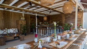 Un lungo tavolo da pranzo in legno apparecchiato con diversi cibi, piatti bianchi e tazze, circondato da sgabelli da bar bianchi e da una panca di legno con cuscini.