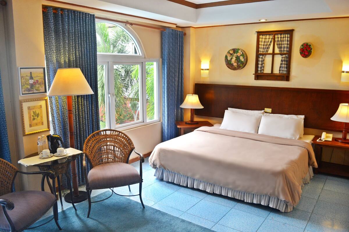 Una habitación de hotel con cama, dos sillas, una mesa con lámpara, una ventana con cortinas y un reloj de pared.