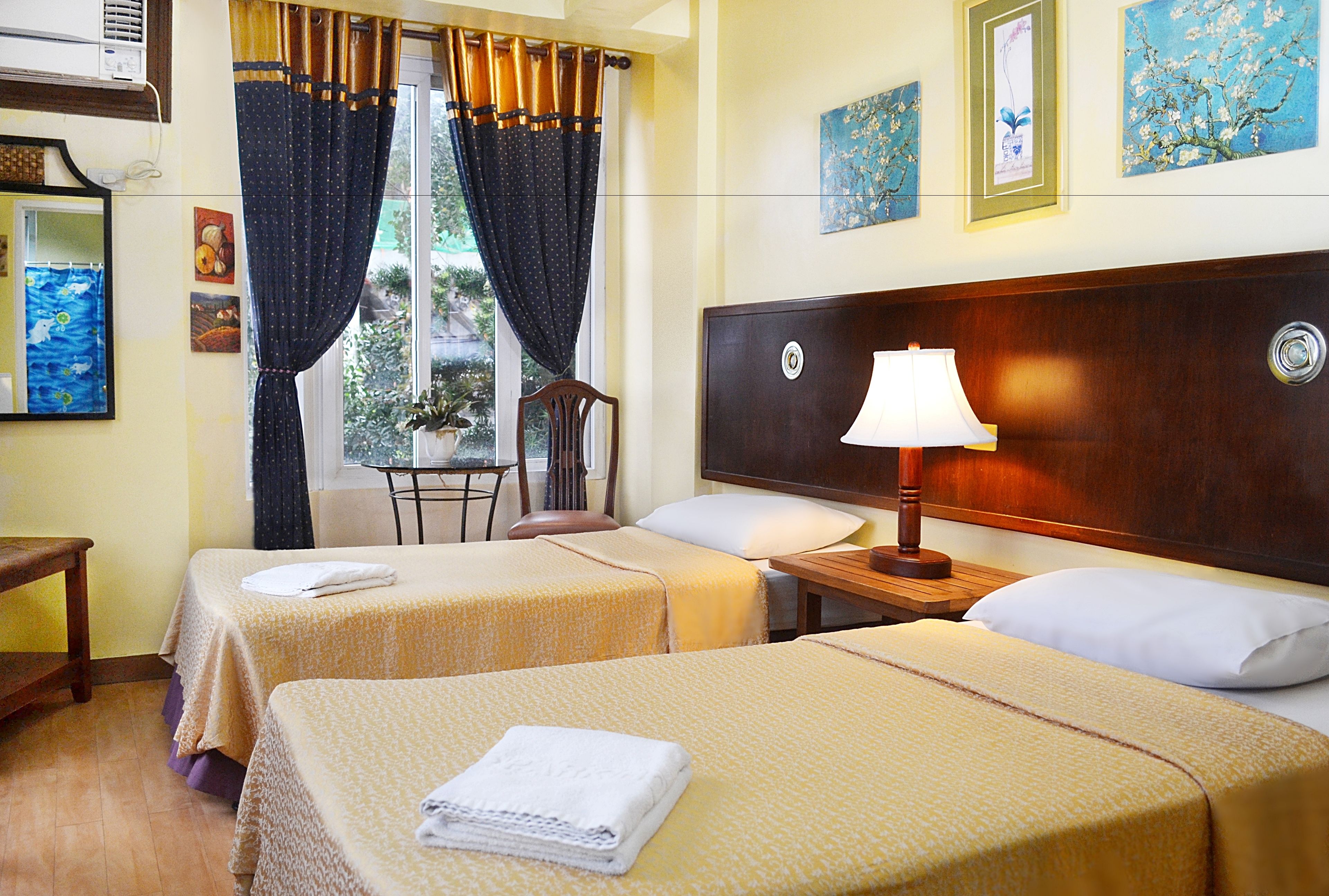 A hotel room with two beds, a wooden headboard, a bedside table with a lamp, a chair, and a window with curtains.