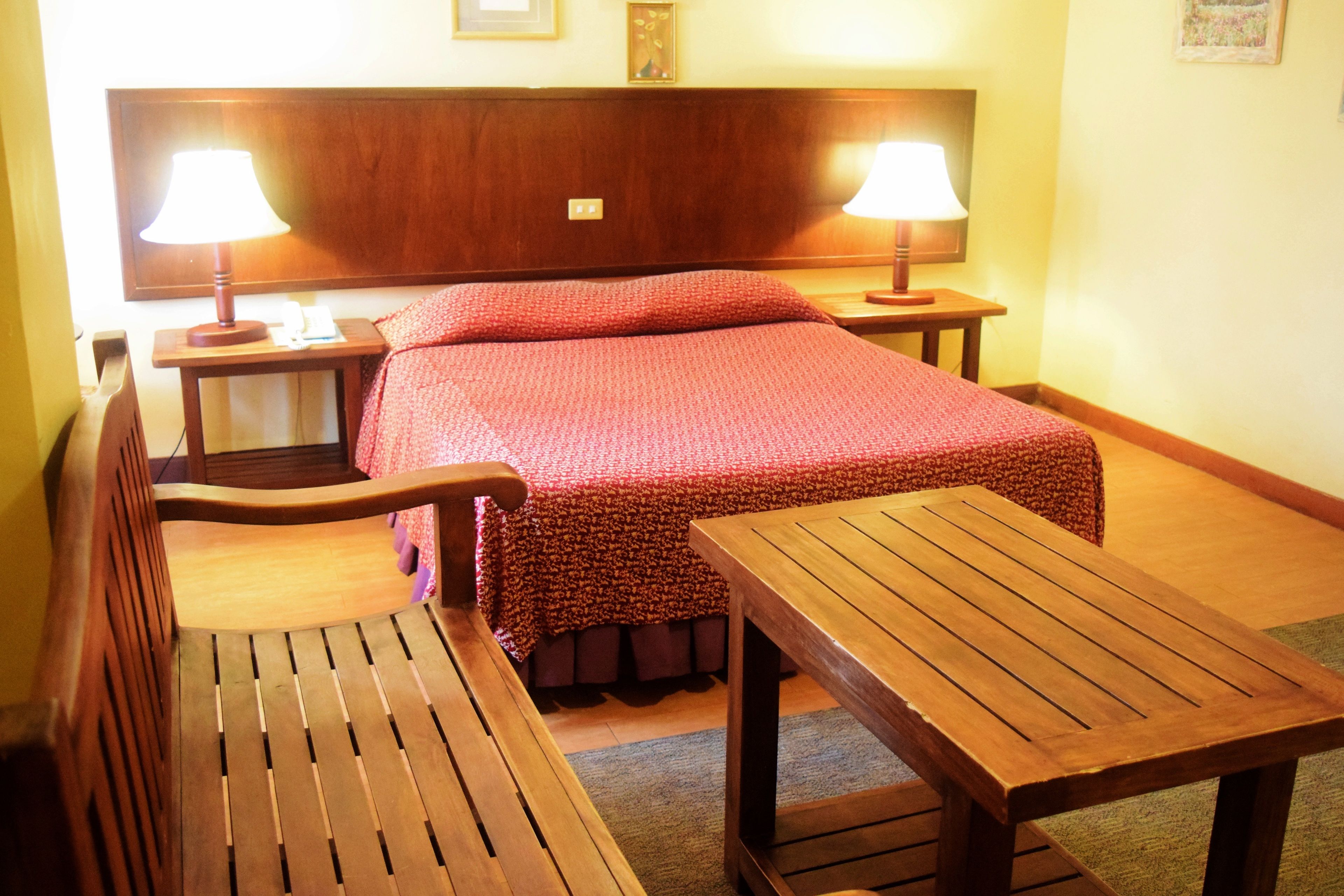A hotel room with a wooden bed, two bedside tables with lamps, a wooden bench, and a small wooden table.