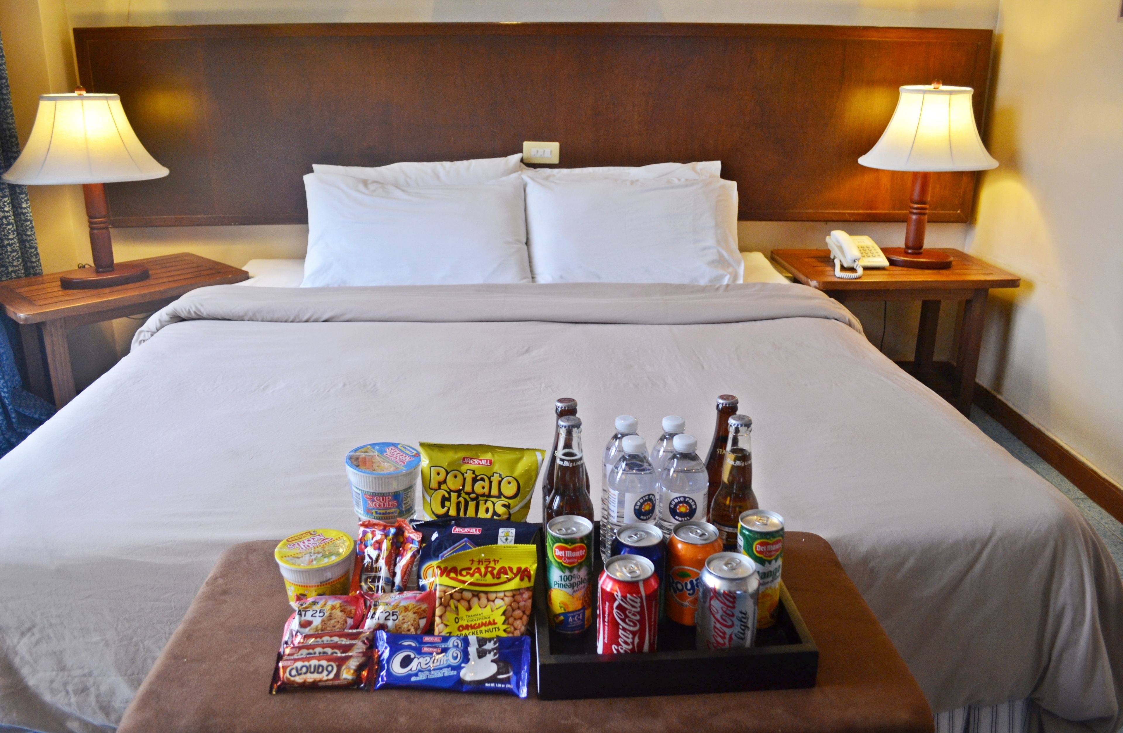 Une chambre d’hôtel avec un lit soigneusement fait, deux lampes de chevet, une petite table avec des collations et des boissons, et un téléphone sur la table de chevet.