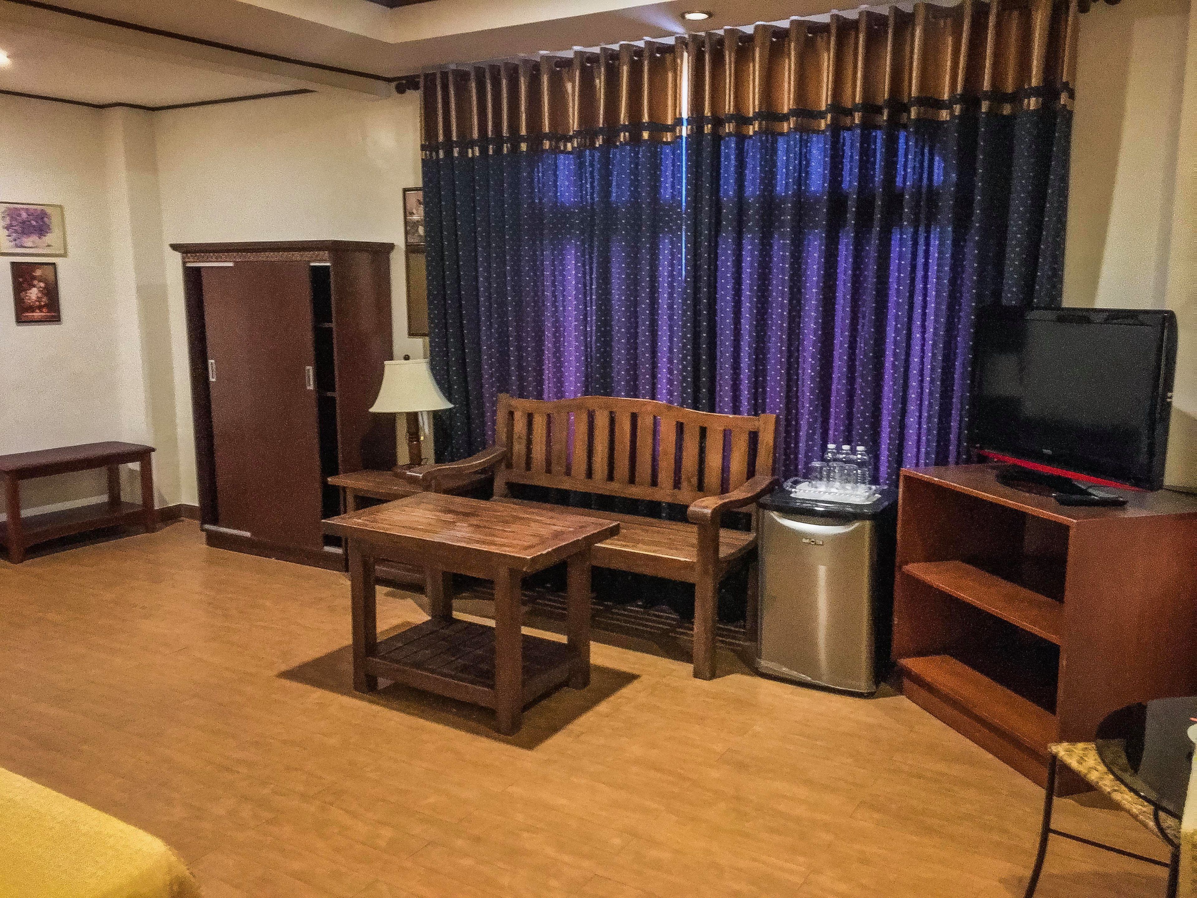 Una habitación con sofá de madera, una mesita pequeña, televisor sobre un soporte, un cesto de basura y un armario.