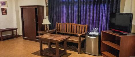 Una habitación con sofá de madera, una mesita pequeña, televisor sobre un soporte, un cesto de basura y un armario.