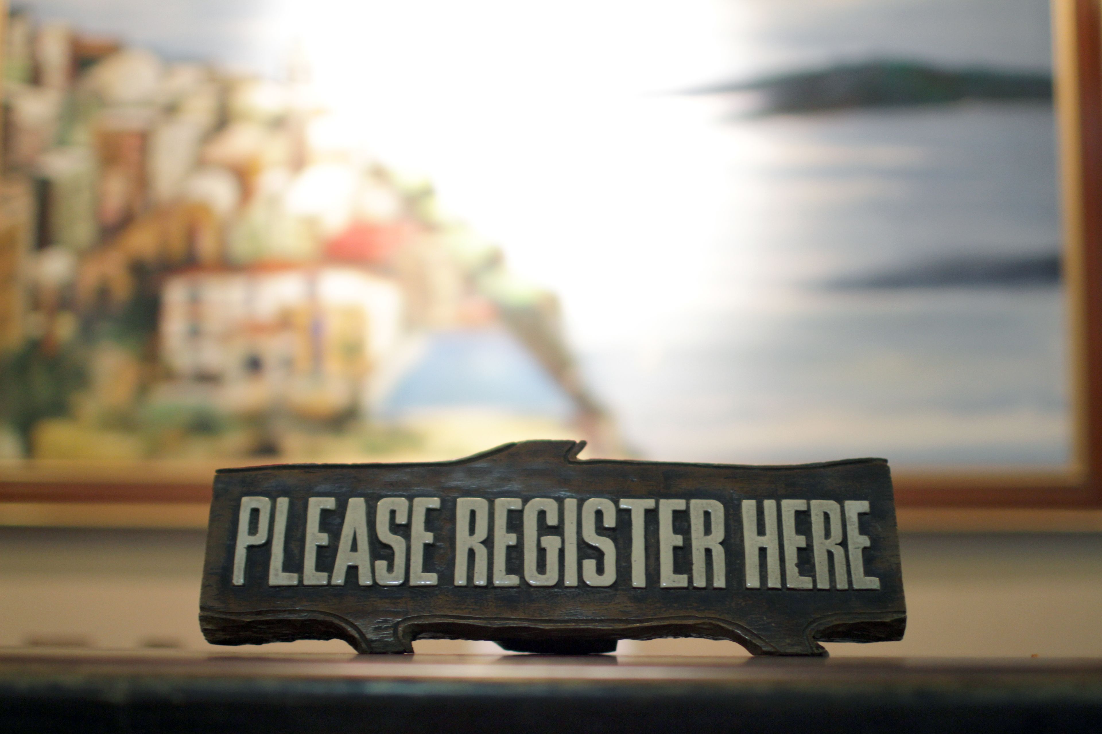 A metal sign with the text "PLEASE REGISTER HERE" in a rustic, weathered style.