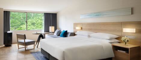 Ein modernes Hotelzimmer mit einem großen Bett, einem Schreibtisch, einem Sessel und Blick ins Freie.