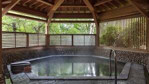 A hot spring area with stone walls and wooden beams, surrounded by a bamboo fence and trees.