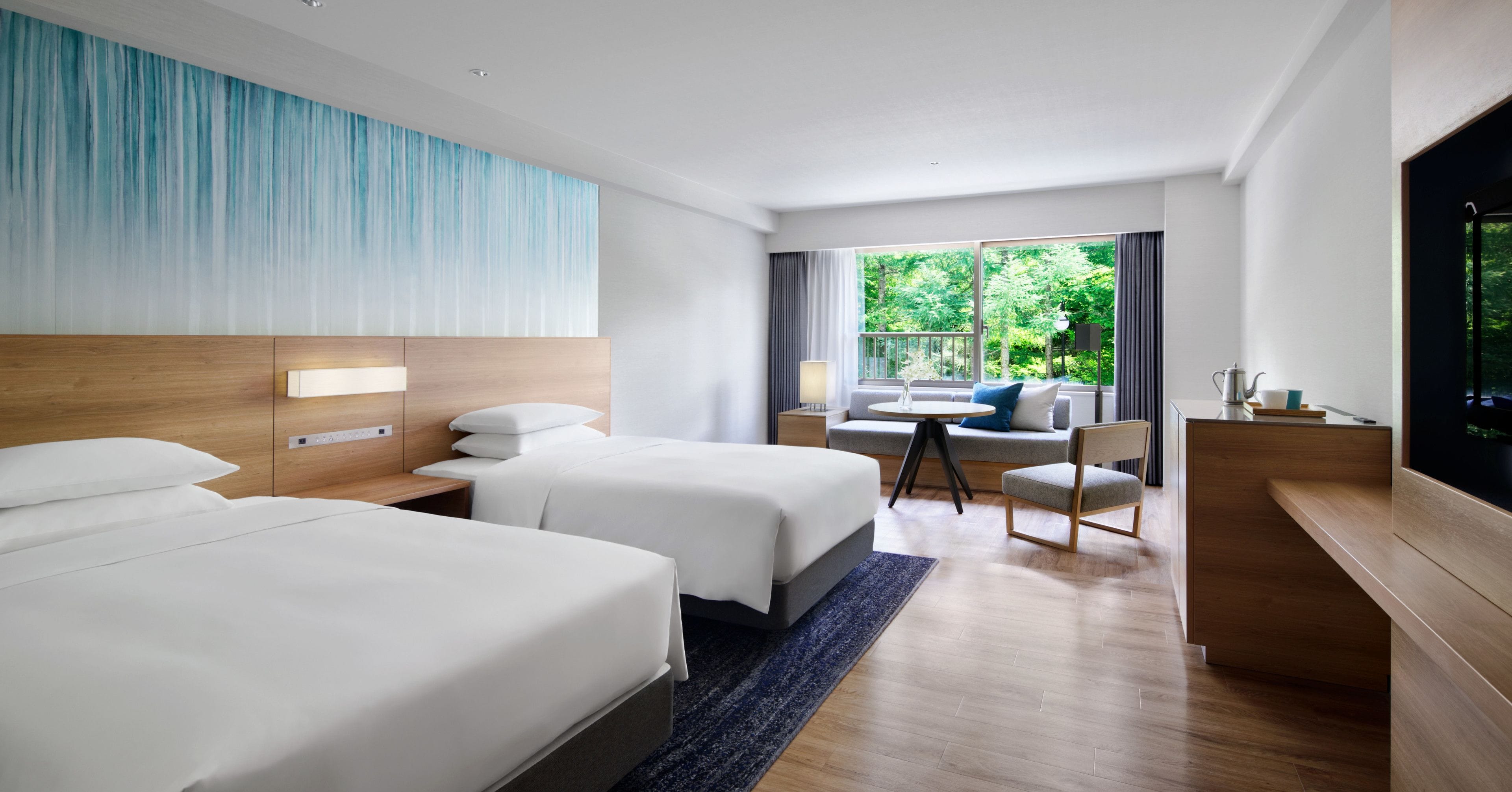 A hotel room with two beds, a desk, a chair, and a large window with a view of greenery.