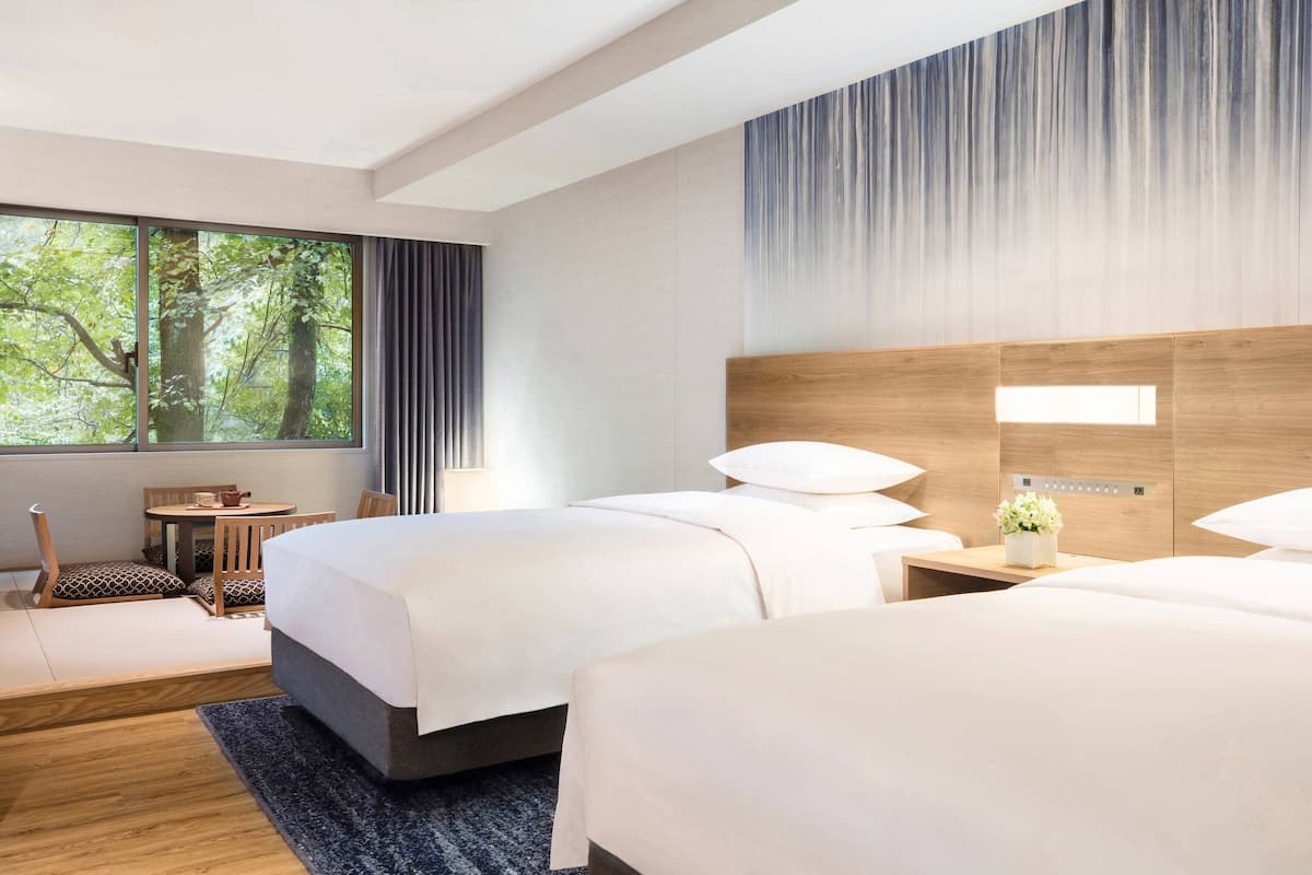 Habitación de hotel con dos camas, una mesita, una silla y ventana con vistas a árboles.