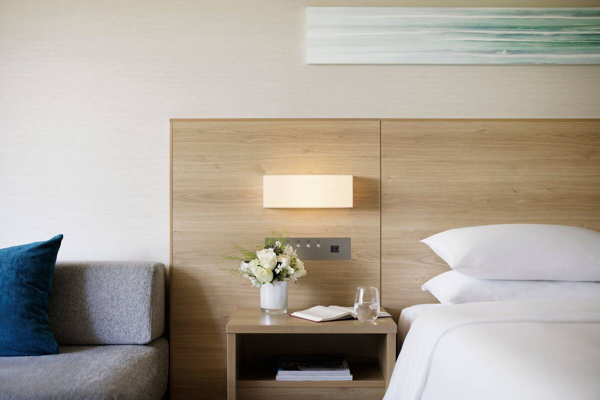 Una habitación de hotel con cabecera de madera, una mesita de noche con un florero, un libro y un vaso de agua.