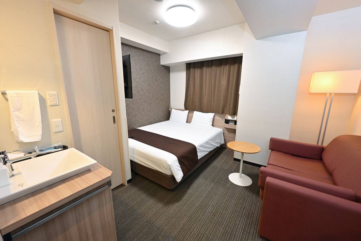 A hotel room with a bed, a small round table, a sofa, a bathroom with a sink, and a mirror.