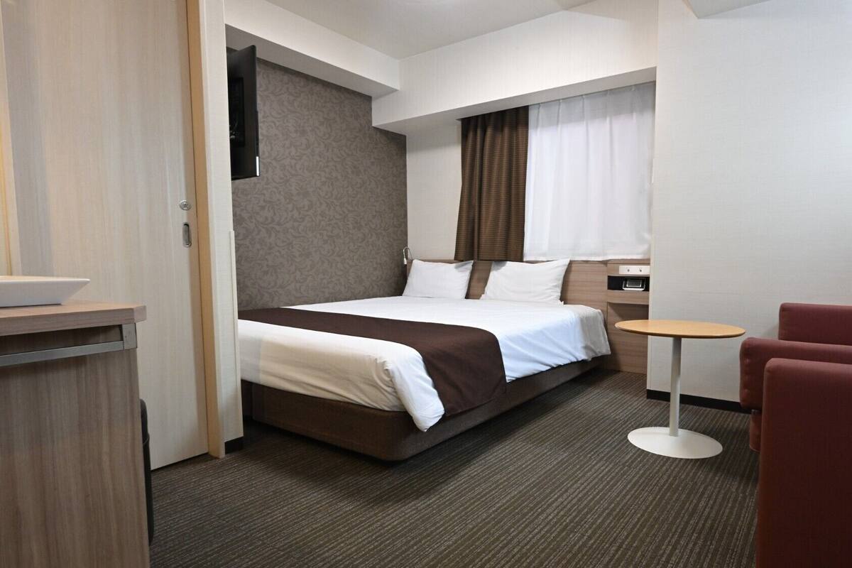 A hotel room with a bed, a small round table, a chair, and a desk with a lamp.