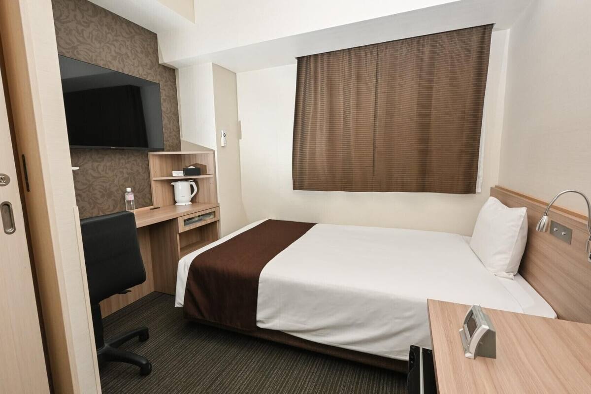 A hotel room with a bed, a desk, a chair, a TV, a coffee maker, and a window with blinds.