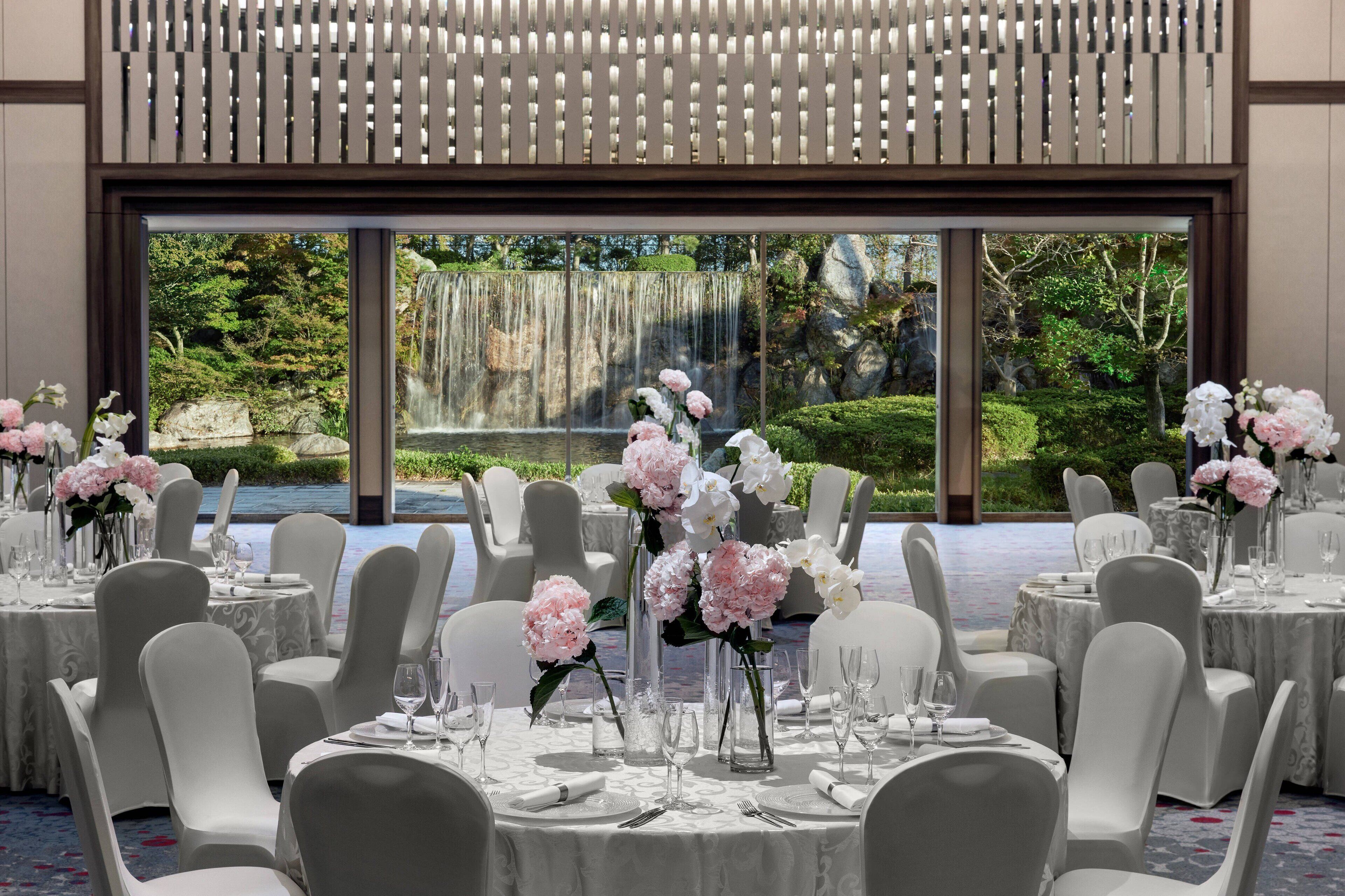 Una sala da banchetti con tavoli rotondi apparecchiati per un evento formale, ampie finestre che offrono vista su una cascata e un giardino.